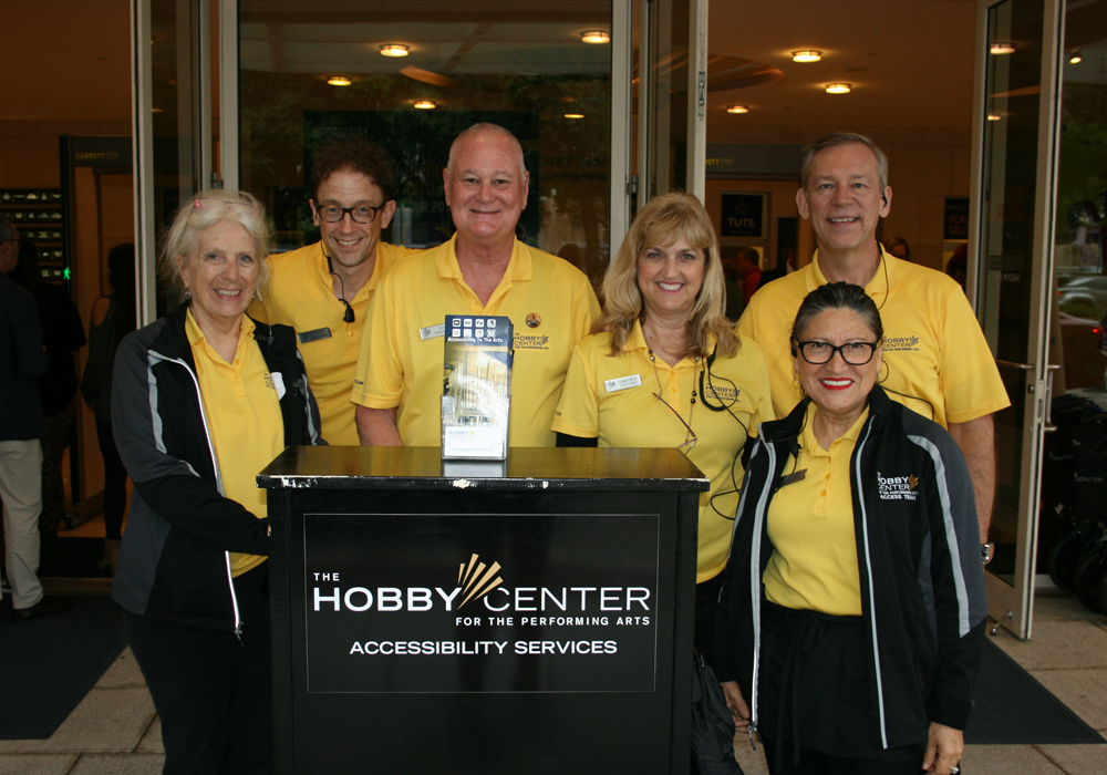 Access team wearing yellow polo shirts gathered around a device and black table with the Hobby Center Accessibility Services Logo on it