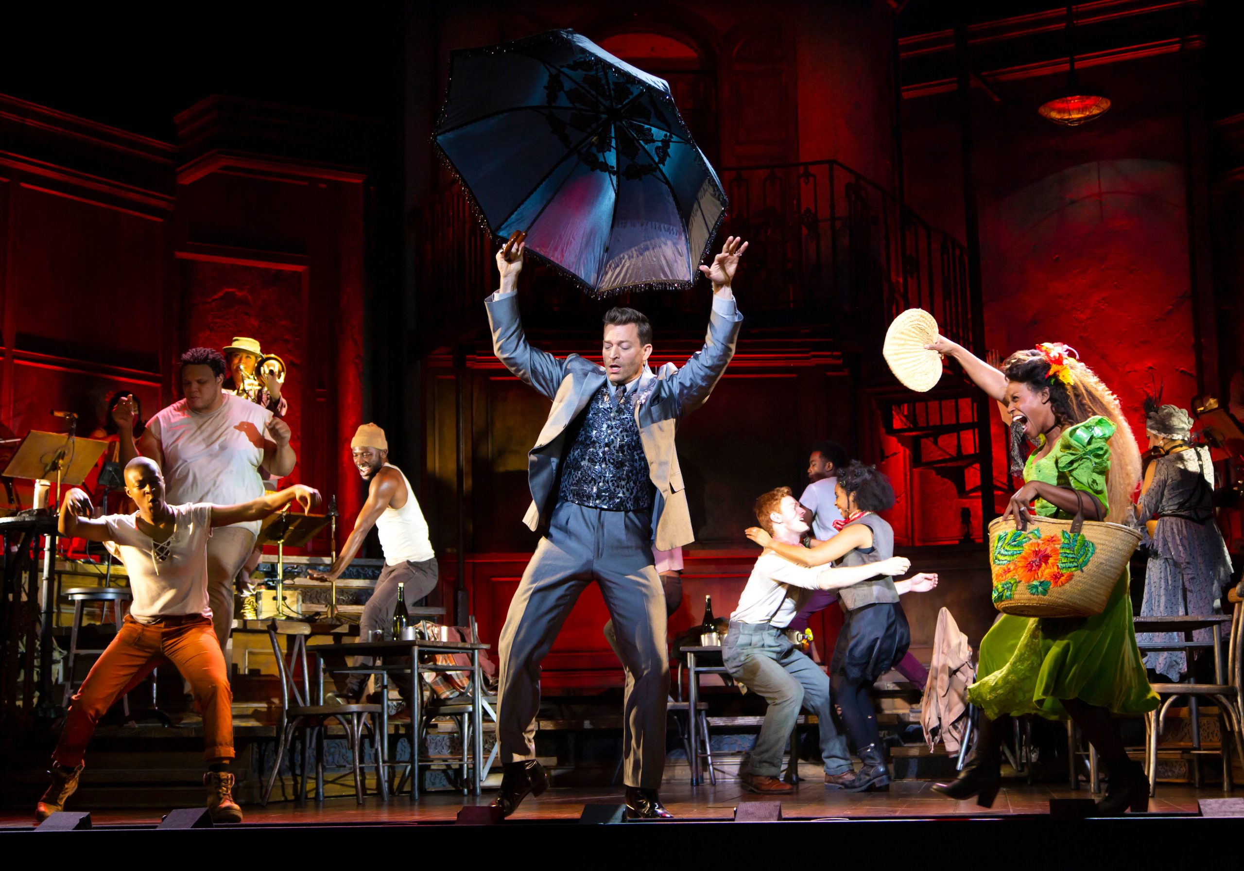 Production image from Hadestown, cast members dance on stage with a man in the center dancing with an umbrella