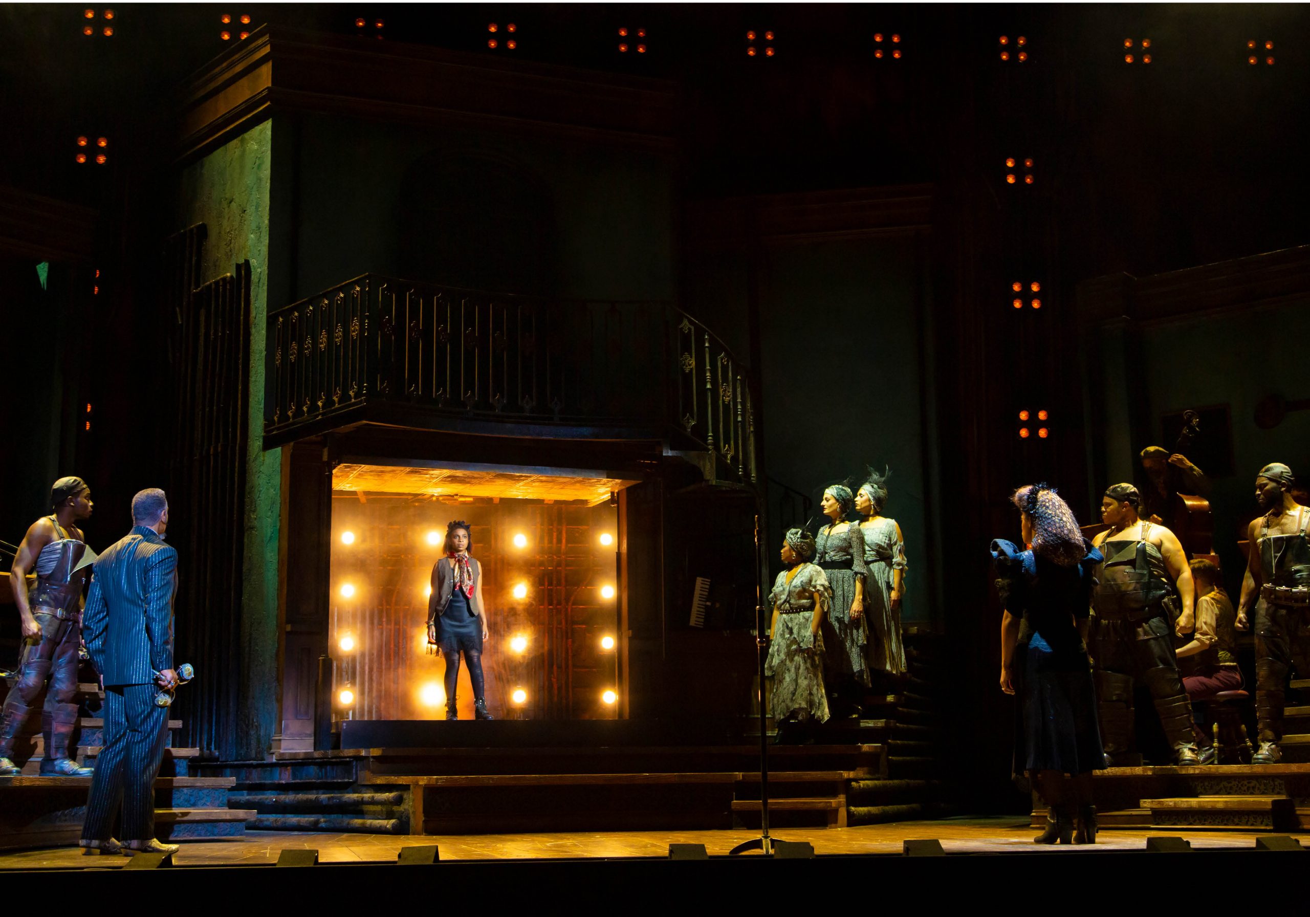 Production image from Hadestown, woman stands on a small stage surrounded by lights and other company members