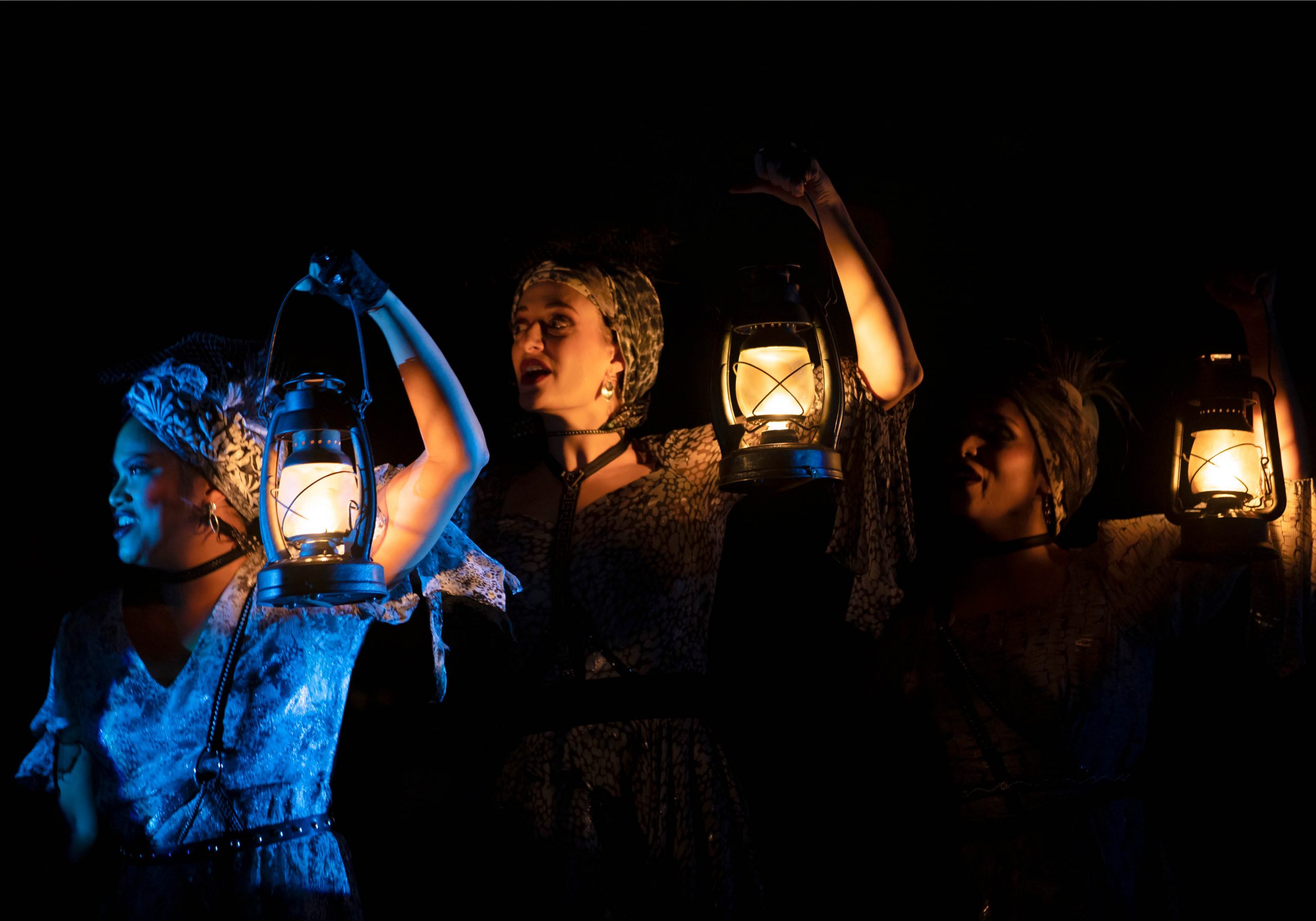 Production image from Hadestown, three women stand in the dark each holding a glowing lantern