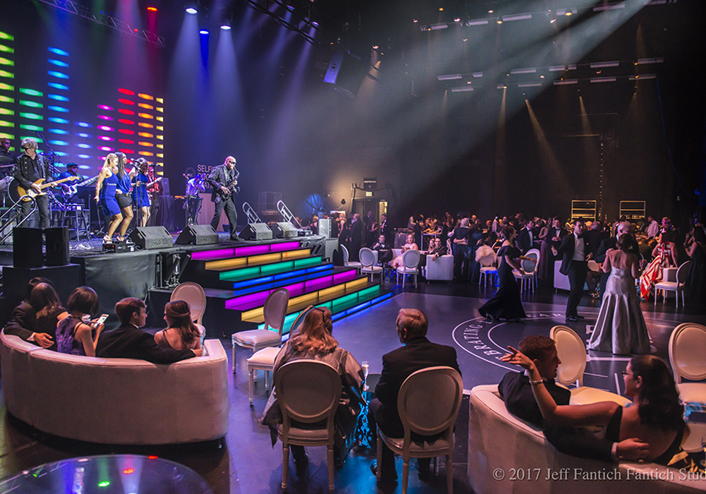 Performance happening on stage with rainbow colored lights in the background and an audience of people watching the performance