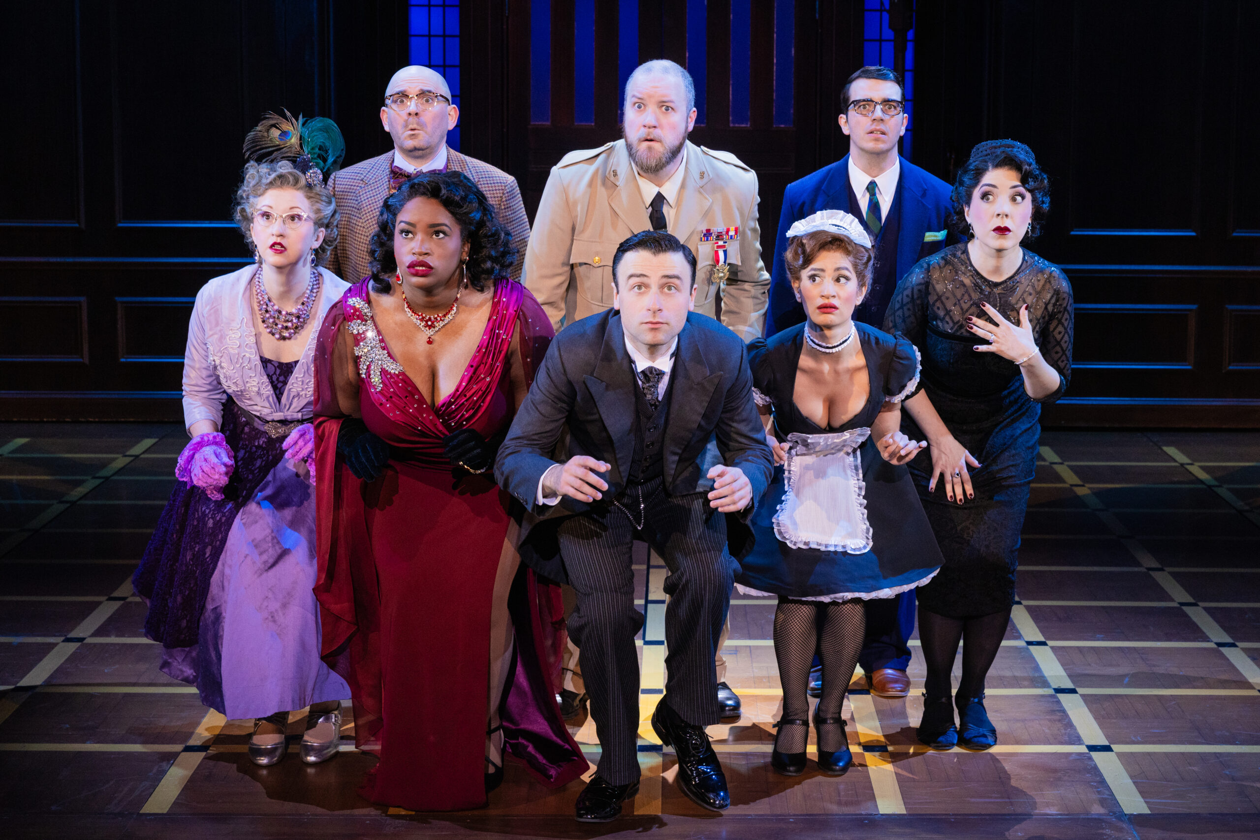 Four women and a man stand on stage crouched in a line with three men standing behind them as they search for something. All actors are looking to their forward off in the distance with a frightened look on their faces.